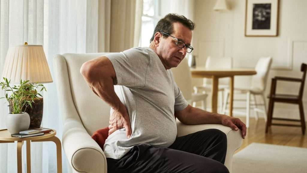 Mann steht mühsam aus einem Sessel auf und hält sich den steifen unteren Rücken wegen Arthrose