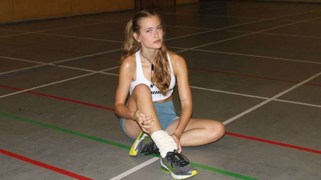 Sportlerin sitzt auf dem Hallenboden und hält ihr umgeknicktes, bandagiertes Sprunggelenk