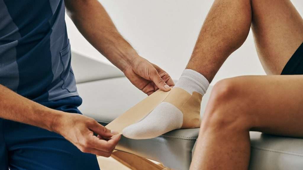 Physiotherapeut legt Sporttape um das Sprunggelenk eines Sportlers auf einer Behandlungsliege